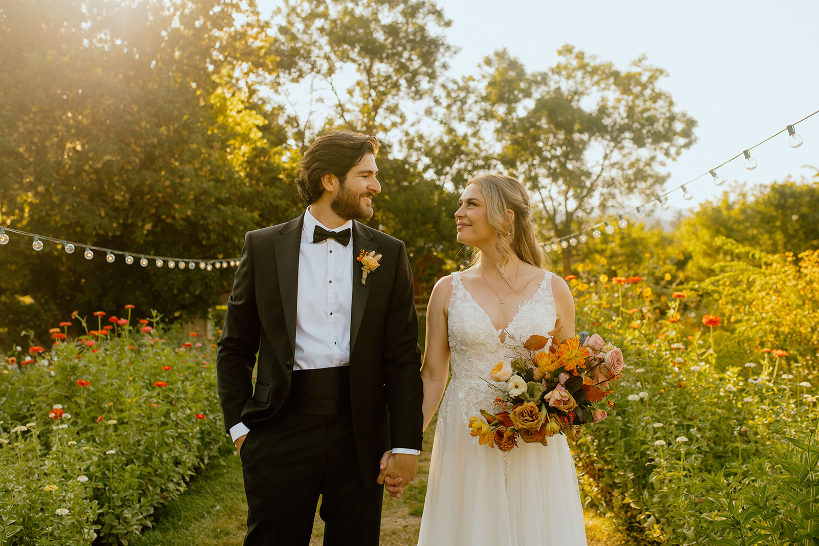 bride and groom smiling at each other holding hands in a flower field
